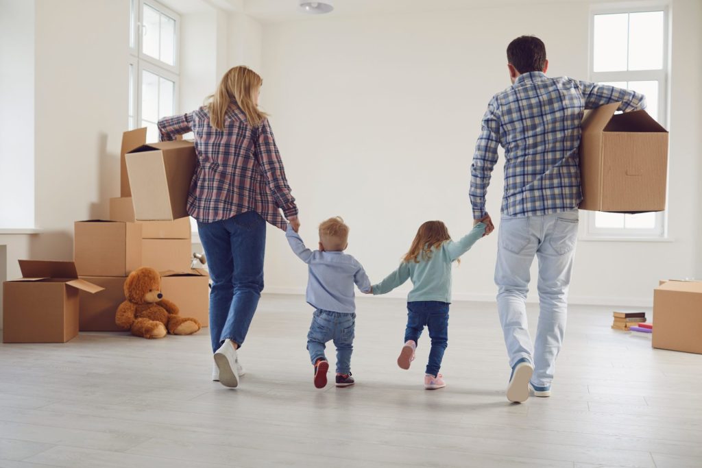  Parents and their two young children hold hands inside their home, moving boxes nearby, preparing to move and manage their own rental property.